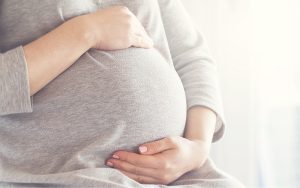 A close-up of a pregnant person gently cradling their belly with both hands, wearing a soft gray top in a bright, softly lit indoor setting.
