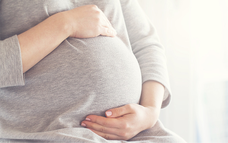 A close-up of a pregnant person gently cradling their belly with both hands, wearing a soft gray top in a bright, softly lit indoor setting.