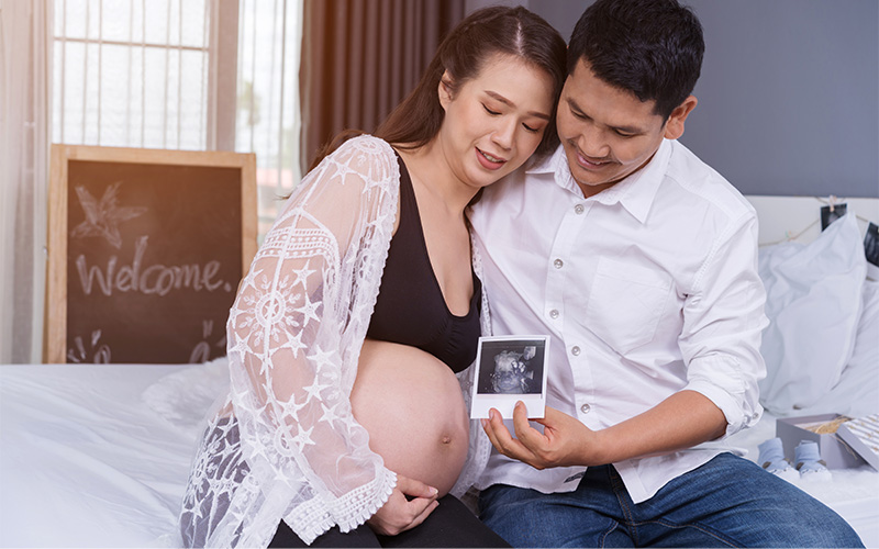 A couple sitting together on a bed, smiling warmly as they look at an ultrasound photo. The pregnant person gently holds their belly while the partner holds the scan, in a cozy, softly lit bedroom with baby-themed decor in the background.