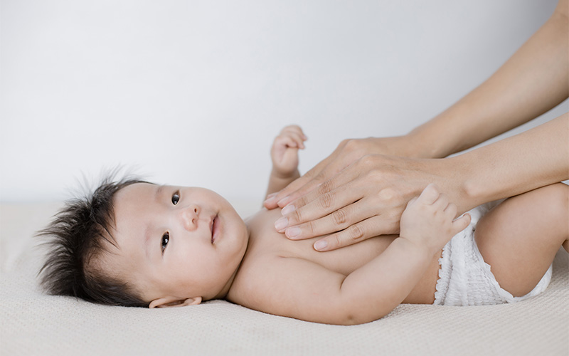 Baby in diaper lying on soft surface with adult hands gently placed on chest in a calm setting.