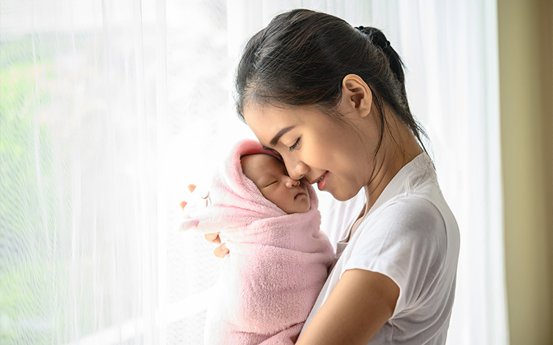 Mother cradling her wrapped newborn baby by window.